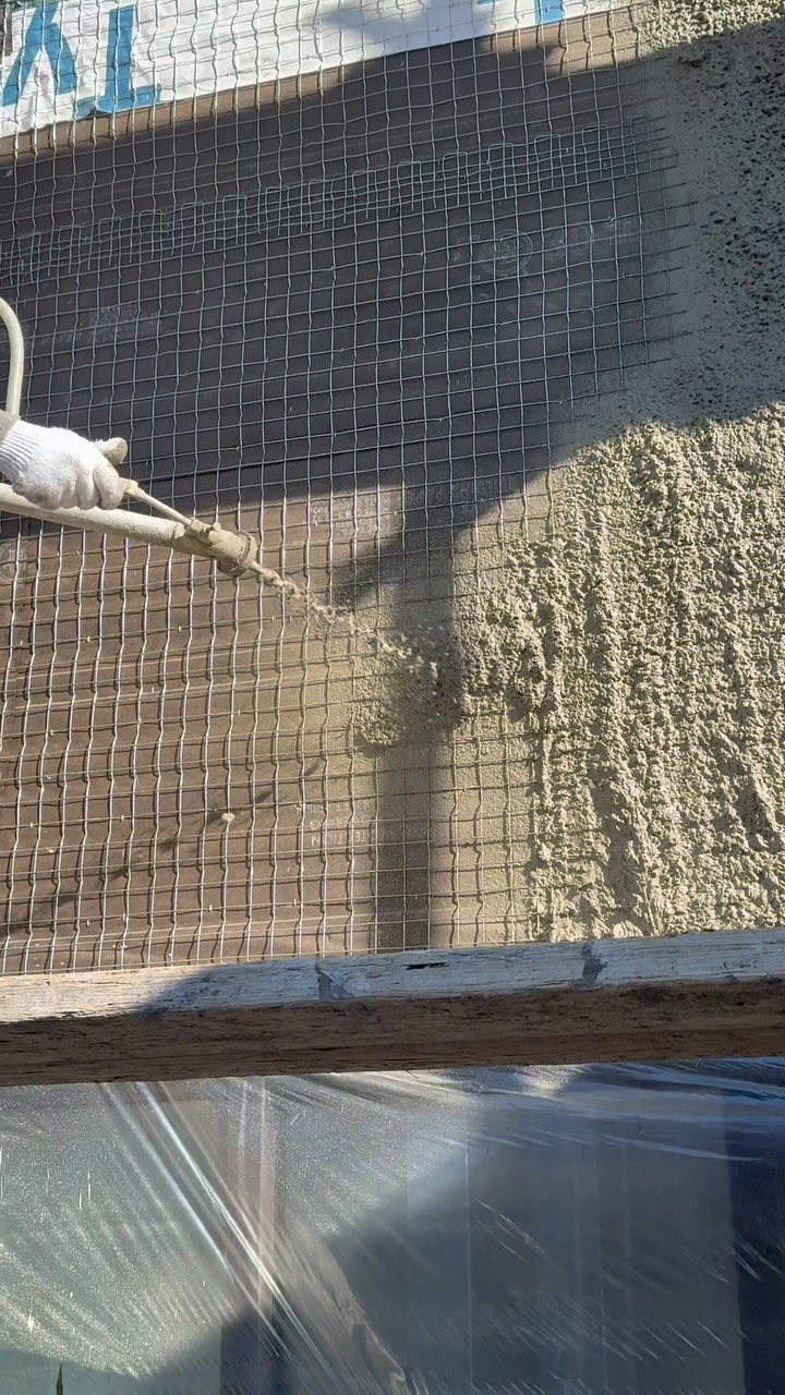 Crew member spray-applying stucco brown coat over galvanized wire with protected windows below